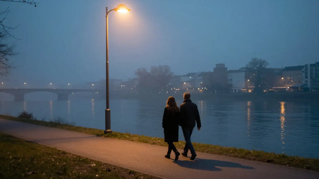 A man and woman walking peacefully along the Spree River at dusk, distant city lights reflecting on water.